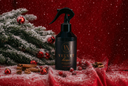 A black spray bottle of Velvet Home on a red fabric surface. Surrounded by cinnamon, berries, red jingle bells, and a snowy branch.