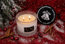 A lit double-wick candle in a frosted glass jar sits on a red fabric surface with scattered faux snow, cinnamon sticks, red berries, and a black lid featuring the brand name Velvet Hollydays.