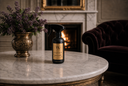 A black spray bottle of Velvet Sparkle Multi-Purpose Spray with a gold label sits on a round white marble coffee table in a luxurious room with a fireplace, a gold vase of purple flowers, and a dark velvet armchair.