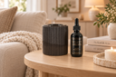 A dark brown ribbed cylindrical container and a black dropper bottle labeled 'VELVET DROPS' sit on a light wood table in a softly lit living room setting with a sofa, books, and a candle.