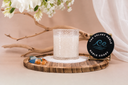 Noble Coast single wick candle atop a pile of sand on a wooden tray. There is also driftwood, florals, sea glass, and amber