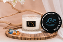 A lit Noble Coast 2 wick candle on a wood tray with sand, sea glass, amber and the lid on the side.