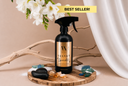 A bottle of Velvet Sparkle Multipurpose Spray on a wooden tray with sea glass, sand, amber and florals on a beige background.