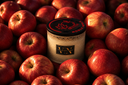 A cylindrical candle jar with a black lid and cream-colored body, surrounded by a dense pile of red apples. The lid features a red maple leaf, cinnamon, and apple logo and the text 'Noble Apple', and the jar has a black label with gold 'VN' lettering.