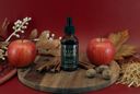 A bottle of Velvet Drops luxury warmer oil centered on a round wooden board, surrounded by red apples, cinnamon sticks, nutmegs, dried leaves, and straw against a deep red background.