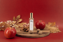 A clear glass bottle with a gold cap and white label reading 'NOBLE APPLE' sits on a round wooden board, styled with a red apple, cinnamon sticks, star anise, whole spices, dried grass, and autumn leaves on a red background.