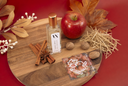 A clear glass bottle with a gold cap and white label reading 'NOBLE APPLE' sits on a round wooden board, styled with a red apple, cinnamon sticks, star anise, whole spices, dried grass, and autumn leaves on a red background.