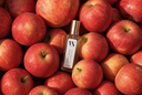 A clear rectangular bottle labeled 'VN' with a gold cap, placed on top of a dense pile of red apples. The bottle label reads 'Noble Apple' and 'Air & Linen Mist'.