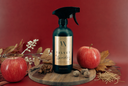 A dark spray bottle labeled 'VELVET Sparkle' stands on a round wooden board, styled with red apples, dried leaves, nuts, and a star anise against a deep red background.
