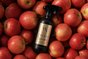 A dark spray bottle labeled 'Velvet Sparkle' surrounded by a dense pile of red apples. The bottle has a black trigger sprayer and a gold label with elegant script text.