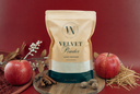 A stand-up pouch of Velvet Powder luxury freshener by The Velvet Noble, styled on a wooden board with red apples, cinnamon sticks, nutmegs, and straw against a deep red background.