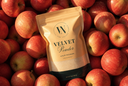 A stand-up pouch of The Velvet Noble Velvet Powder luxury freshener, styled on a bed of red apples. The pouch is a warm beige color with a black base and features the brand's logo and name clearly printed on the front.