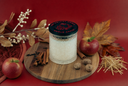 A glass jar candle with a black lid labeled 'The Velvet Noble' and 'Noble Apple' sits on a round wooden board, surrounded by apples, cinnamon sticks, straw, and autumn leaves on a red background.
