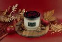 A jar candle labeled 'VN' sits on a round wooden board, surrounded by autumn props including red apples, cinnamon sticks, dried leaves, and straw against a deep red background.