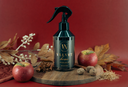 A dark spray bottle labeled 'VELVET home' stands on a round wooden board with apples, cinnamon sticks, star anise, and dried leaves against a deep red background.