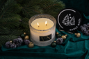 A lit double-wick candle in a textured clear glass jar sits on dark teal fabric, surrounded by pine branches, purple berries, gold and green ornaments, and a round black coaster printed with the brand name NOBLE NOÉL.