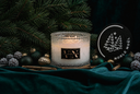A lit scented candle in a textured clear glass jar sits on a dark teal velvet surface, surrounded by pine branches, holiday ornaments, and a round black lid printed with a tree logo and the words 'NOBLE NOEL'.