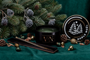 A black scented candle in a matte black jar with gold lettering, styled with pine branches, pinecones, and holiday ornaments on a dark green fabric background. A round lid with a white Christmas tree graphic and the text 'NOBLE NOËl' is visible on the right, and a gold-toned decorative rod rests in an open black box on the left.