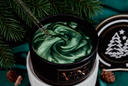 A close-up of a black candle jar filled with a swirled green wax, set on a white pedestal with pine branches, a gold glitter dropper, and a lid featuring a white Christmas tree design.