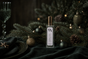 A clear rectangular bottle with a gold cap and a white label reading 'VN Noble Noel' stands on dark green fabric surrounded by pine branches, ornaments, and a glass in a moody holiday setting.