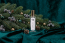 A clear rectangular bottle with a gold cap and a white label reading 'Noble Noel' and 'Air & Linen Mist', styled on dark teal velvet with pine branches, frosted berries, pinecones, and gold ornaments.