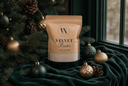 A stand-up pouch of Velvet Powder luxury freshener by The Velvet Noble, styled on a dark green velvet cloth with Christmas ornaments and a decorated Christmas tree in the background.