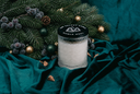 A frosted glass candle jar with a black lid printed with a white mountain logo and the words 'The Velvet Noble Noble Noel,' styled on dark teal fabric with pine branches, berries, and gold ornaments.