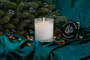 A lit white candle in a textured clear glass holder sits on draped teal velvet with pine branches, pinecones, and holiday ornaments around it. A round black sign reads 'THE VELVET NOBLE NOBLE NOEL' with a tree illustration.