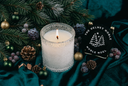A lit white candle in a textured clear glass holder sits on dark teal fabric surrounded by pine branches, pinecones, berries, and ornaments. A black hanging tag with white text reading 'THE VELVET NOBLE NOBLE NOEL' and a tree graphic is placed beside the candle.