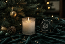 A lit frosted glass candle sits on dark green velvet next to a round black sign reading 'THE VELVET NOBLE / NOBLE NOEL' with a tree illustration, in front of a decorated Christmas tree with warm lights.
