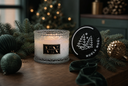 A lit scented candle in a textured clear glass jar sits on a wooden surface surrounded by pine branches, green and gold ornaments, and a round black lid with white tree graphics and the words 'NOBLE NOEL'. A blurred Christmas tree with warm lights appears in the background.