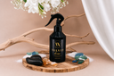 A bottle of Velvet Home Air & Linen Mist on a wooden tray with sea glass, sand, amber and florals on a beige background.