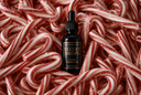 A small black dropper bottle labeled 'Velvet Drops Luxury Warmers Oil' stands centered on a dense arrangement of red-and-white striped candy canes with fine sugar dusting.