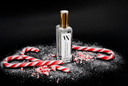A clear glass perfume bottle with a gold cap and a white label reading 'VN' and 'PEPPERMINT LEGACY', styled on a black background with red-and-white candy canes and crushed candy cane pieces.