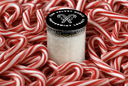 A small clear glass jar filled with white peppermint candy, topped with a black lid printed with 'THE VELVET NOBLE PEPPERMINT LEGACY,' surrounded by a dense bed of red-and-white striped candy canes. The jar is centered and the lid text is legible.