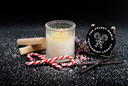 A frosted glass candle jar with white wax sits on a dark textured surface with red-and-white candy canes, vanilla beans, pink peppermint pieces, and a round black sign reading 'The Velvet Noble Peppermint Legacy.'.