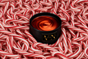 A black cylindrical candle jar with a gold VN logo sits in the center of a dense pile of red and white striped candy canes. The candle wax inside is a warm red  with a visible swirl pattern.