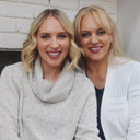 Two women with blonde hair sitting together against a white brick wall.