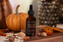 An amber glass bottle of Velvet Drops Luxury Warmer Oil on a wooden surface with decorative fall elements.