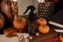 A dark black velvet home fragrance spray bottle with a black trigger sprayer, styled on a wooden board with miniature pumpkins, autumn leaves, acorns, and a candlestick.