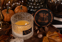 A lit scented candle in a textured glass jar sits on a rustic wooden surface surrounded by pumpkins, autumn leaves, books, and a black round lid with a pumpkin illustration. The candle label reads 'VN' and 'Velvet Pumpkin' is visible on the lid.