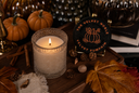 A lit candle in a textured glass holder sits on a wooden tray beside a round black coaster printed with the words 'The Velvet Noble Velvet Pumpkin' and an orange pumpkin illustration. The scene is styled with autumn leaves, pinecones, cinnamon sticks, and small pumpkins in a warm, dark setting.