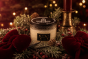 A holiday-themed jar candle from The Velvet Noble, styled with red velvet bows, pinecones, and evergreen branches on a dark festive background with warm bokeh lights.