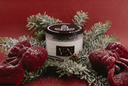 A white candle in a clear glass jar with a black lid and a black label reading VN The Velvet Noble, styled with red velvet bows, green frosted pine branches, and a cinnamon stick on a deep red background.