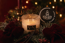 A lit candle in a textured glass jar sits on a holiday arrangement with greenery, red velvet bows, berries, a brass candlestick, and a round black label reading 'SILENT NIGHT'.