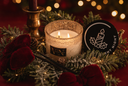 A lit candle in a textured clear glass holder sits on a red surface surrounded by green pine branches, red velvet bows, and warm string lights. A round black product tag with a candle illustration and the words 'SILENT NIGHT' is visible beside the candle.