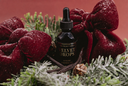 A small black dropper bottle labeled 'The Velvet Noble Velvet Drops' centered among red velvet bows, frosted greenery, and a warm red background.