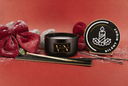 A black candle jar with a gold logo plaque sits on a red surface beside a round black lid labeled 'SILENT NIGHT' and a decorative red bow with silver pine branches.