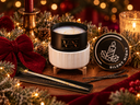 A holiday-themed scented candle set displayed on a wooden surface with pine branches, red velvet fabric, and warm bokeh lights. The main product is a black tin candle in a white ribbed ceramic holder, with its lid placed beside it.