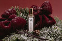 A clear rectangular perfume bottle with a gold cap and a white label reading 'Silent Night' stands centered in a holiday wreath made of dark red velvet bows, green frosted pine branches, vanilla beans, and a small pinecone against a deep red background.
